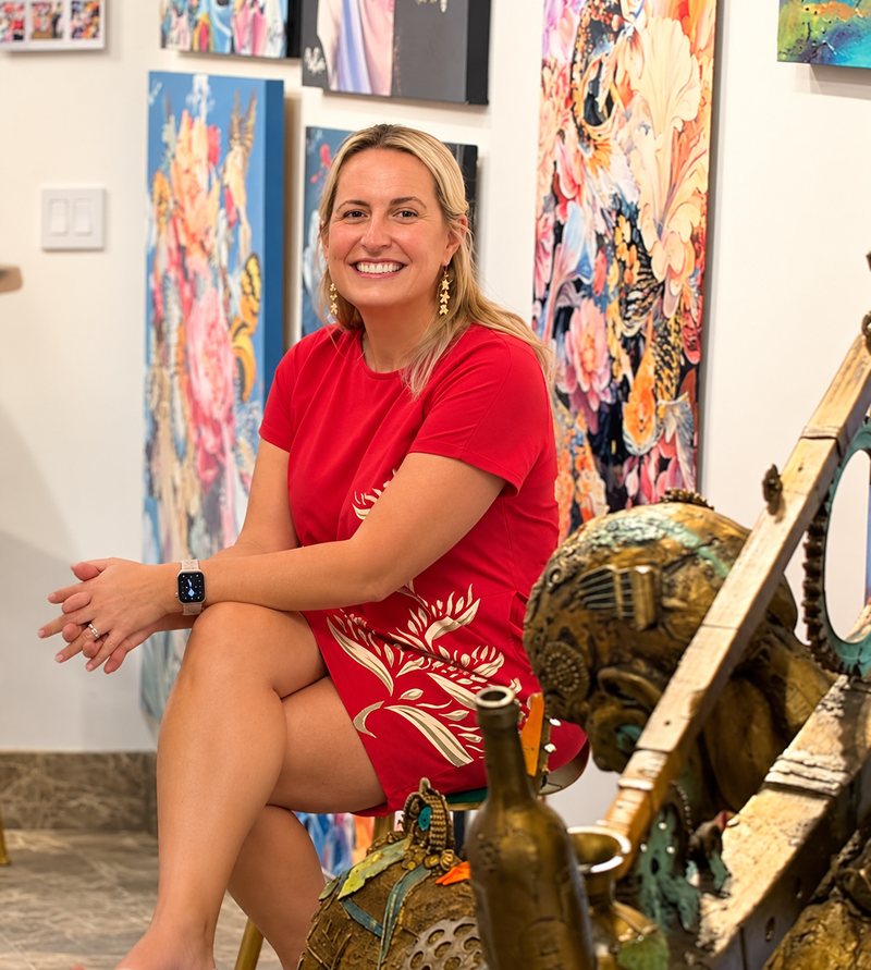 Dr. G smiling, seated in a red dress among colorful floral paintings at the gallery